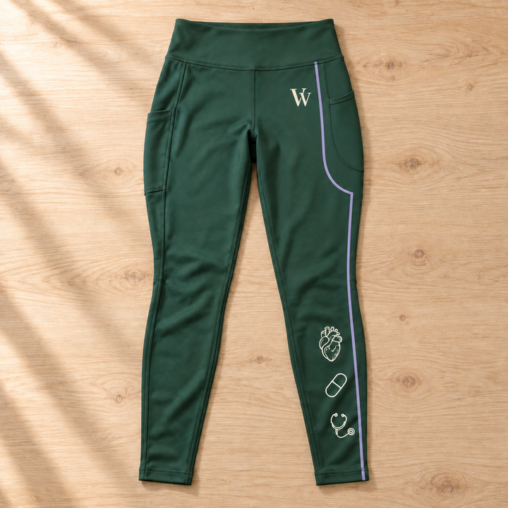 Forest-green pocket leggings laid flat on a wooden table, showing the cream VV, medical illustrations and lavender stripe following the thigh pocket contour.