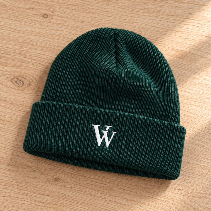 Spruce cuffed beanie laid naturally on pale oak, showing the ribbed knit, folded cuff and white VV embroidery.