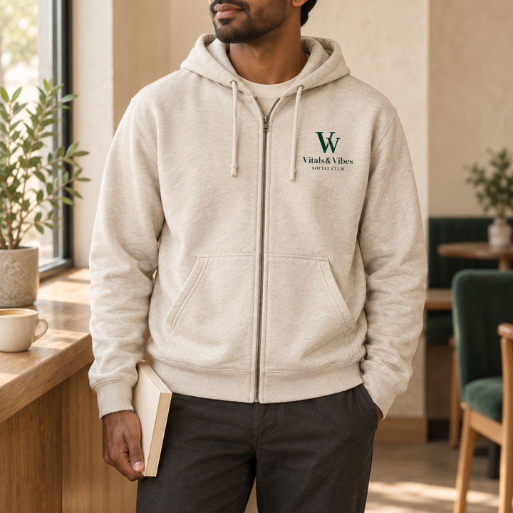 Person wearing an oatmeal full-zip hoodie with a green VV, Vitals & Vibes and Social Club chest logo beside a cafe window.