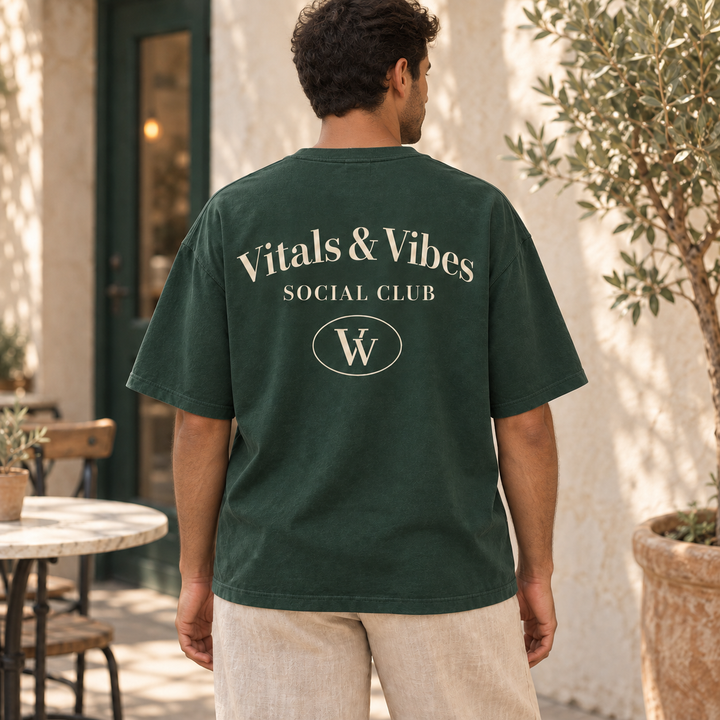 Man seen from behind wearing a forest-green oversized tee with cream Vitals & Vibes, Social Club and an oval VV emblem in a courtyard.
