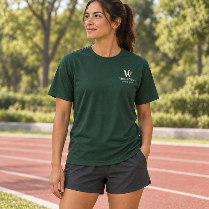 Woman wearing a forest-green performance tee with a small cream VV and Social Club chest logo beside a running track.