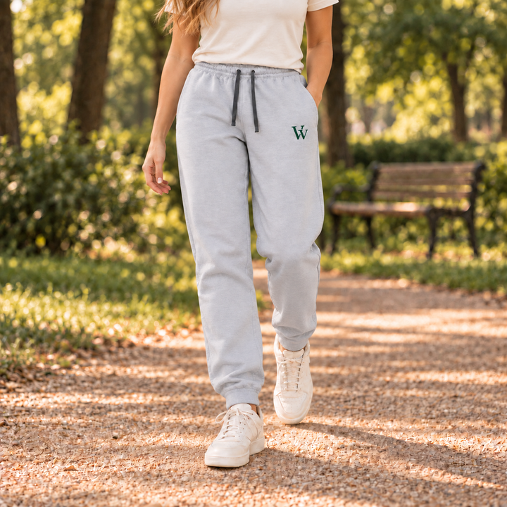 Woman walking on a park path in Athletic Heather joggers with dark drawcords and a small green VV embroidery on the left thigh.