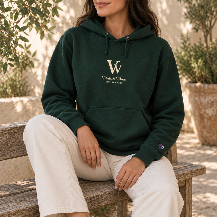 Woman seated on a wooden bench in a sunny courtyard wearing a dark green VV Social Club Champion hoodie.