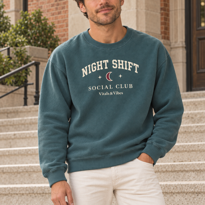 Man standing on pale stone steps in a Blue Spruce Night Shift Social Club crewneck and cream trousers.