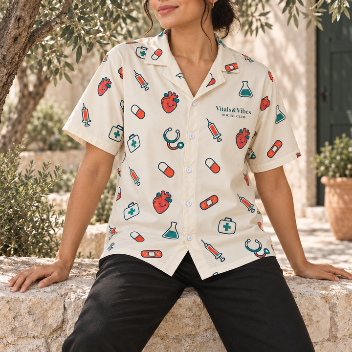 Woman seated on a pale stone wall beneath an olive tree wearing the cream medical scatter button shirt.