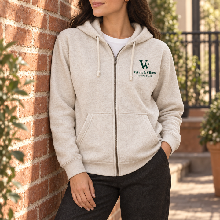 Woman leaning against a brick courtyard wall wearing an oatmeal full-zip VV Social Club hoodie.