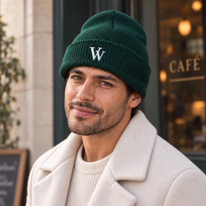 Man wearing a spruce-green VV cuffed beanie in a warmly lit café, with the white embroidered monogram in clear view.